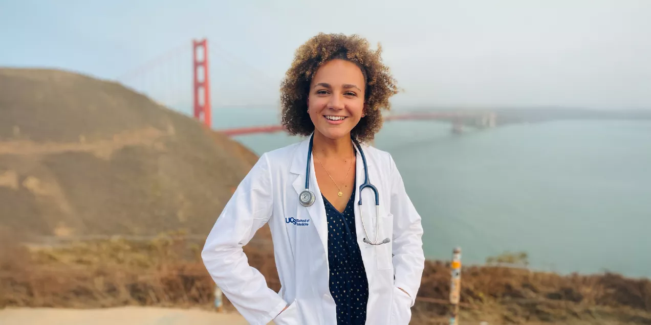 Portrait of a smiling person with curly hair wearing a white UCSF School of Medicine lab coat and a stethoscope over a dark patterned top. They are standing outdoors with the Golden Gate Bridge and San Francisco Bay softly blurred in the background.