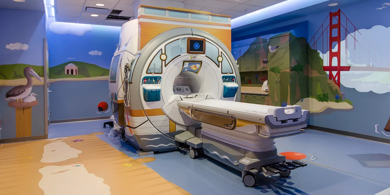 A brightly decorated pediatric MRI suite with a colorful beach and San Francisco-themed mural surrounding the scanner. The MRI machine is designed with child-friendly artwork, and the room features playful illustrations to create a welcoming environment for young patients.