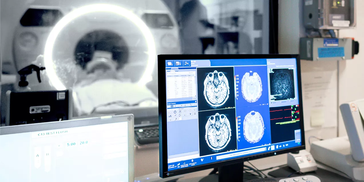 A radiology workstation displays brain MRI scans and imaging data as a patient is positioned inside an MRI scanner visible through a control room window.