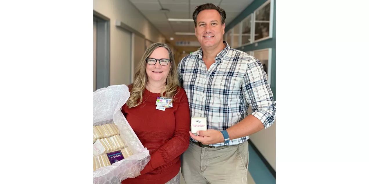 Two individuals stand together in a hospital hallway smiling toward the camera. One person holds a decorated box containing individually wrapped treats, while the other holds a small card related to MRI Safety Week. Both wear identification badges, and the hallway is softly lit with clinical and administrative spaces visible in the background.
