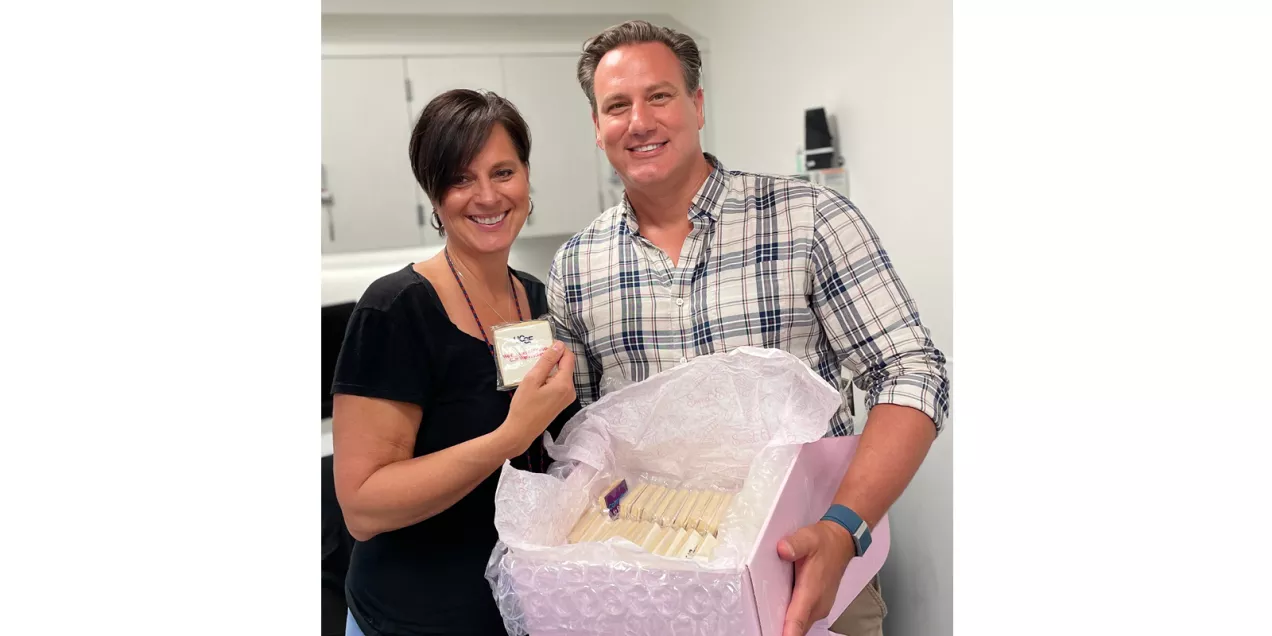 Two individuals smile for a photo while holding a box of individually wrapped treats during an MRI Safety Week activity. One person displays a small card, and both stand in a clinical workspace with cabinets and equipment visible in the background.