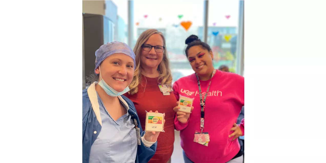 Three healthcare workers pose together in a clinical setting during MRI Safety Week. Two individuals hold safety-themed cards while smiling toward the camera. One person is dressed in surgical attire with a mask lowered beneath the chin, while the others wear hospital identification badges. Large windows and colorful decorations are visible in the background.