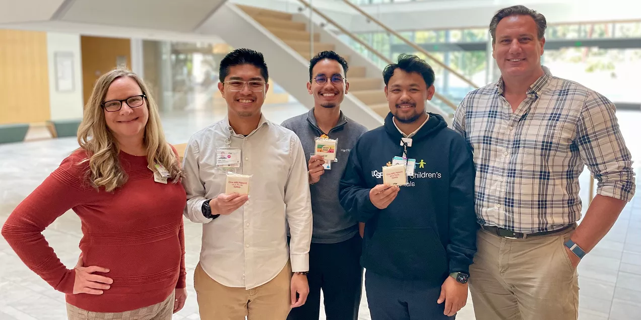 Five individuals stand together in a bright, modern lobby holding small cards or badges and smiling toward the camera. The group is casually dressed, with one person wearing a UCSF Benioff Children's Hospitals sweatshirt. A staircase and large windows are visible in the background, creating an open and welcoming environment. The photo appears to have been taken during a workplace event, training activity, or recognition program.