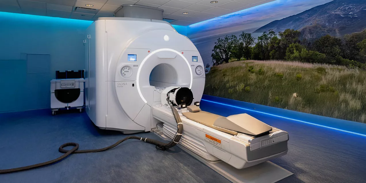 Wide view of an MRI scanner with the patient table extended in a modern imaging suite. The room features blue accent lighting and a large wall mural of a grassy landscape with trees and mountains, creating a calming environment.