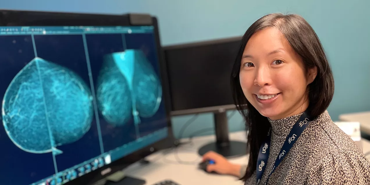 A smiling person sits at a computer workstation, looking toward the camera while viewing mammography images displayed on a large monitor. They are wearing a patterned long sleeve top and a UCSF lanyard in a clinical imaging office.