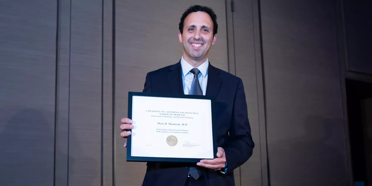 Mark Mamlouk stands in a suit and tie, smiling while holding a framed UCSF certificate recognizing his service as a volunteer physician. He is photographed indoors against a neutral background during an awards ceremony.
