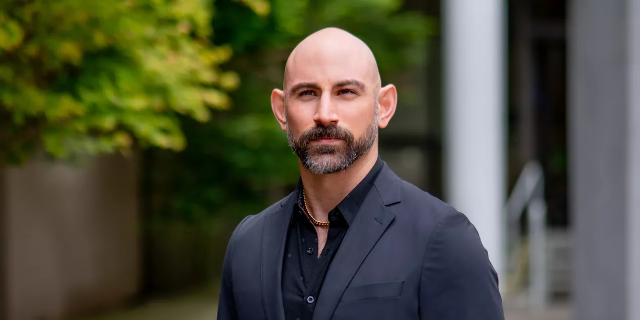 Professional portrait of a man with a shaved head and neatly trimmed beard wearing a dark suit jacket over a black shirt. He stands outdoors with a serious expression, while greenery and modern building elements are softly blurred in the background.