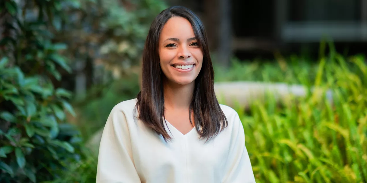 Smiling woman with shoulder-length dark hair wearing a cream-colored blouse, standing outdoors in front of softly blurred greenery and landscaping. She is facing the camera with a friendly expression in a natural garden setting.