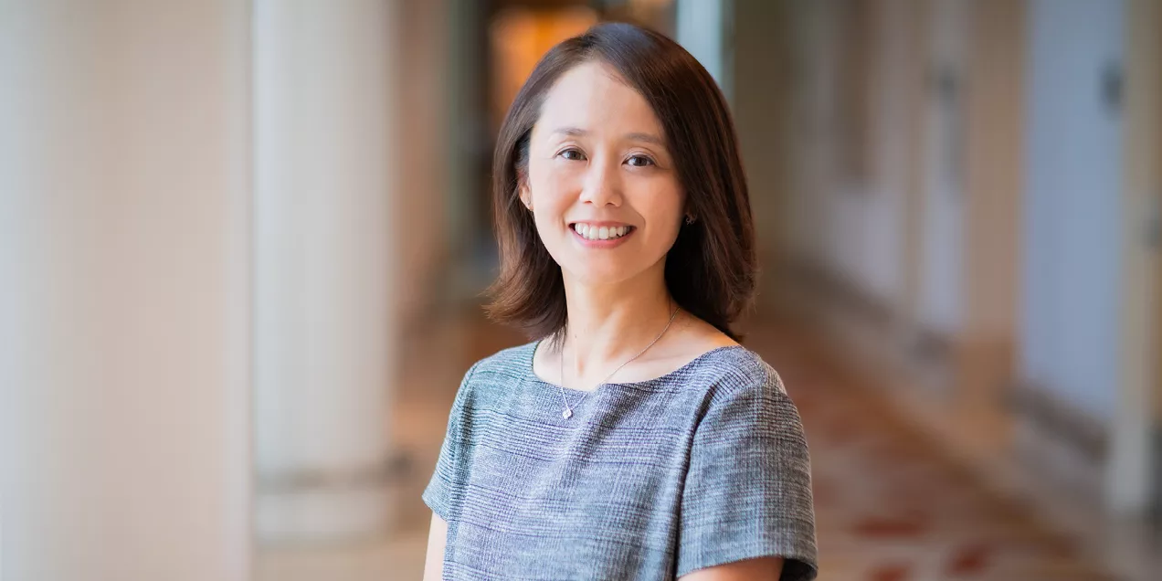 Professional portrait of a woman with shoulder-length dark hair wearing a gray short-sleeved dress and a delicate necklace. She is smiling at the camera while standing in a softly lit indoor hallway, with columns and a patterned floor blurred in the background.