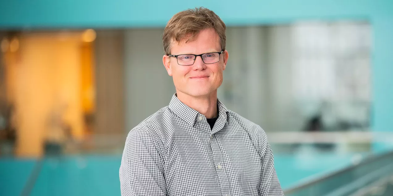 Portrait of Peder Larson wearing glasses and a checkered button-down shirt, smiling at the camera. He stands indoors with a softly blurred modern building interior in the background.