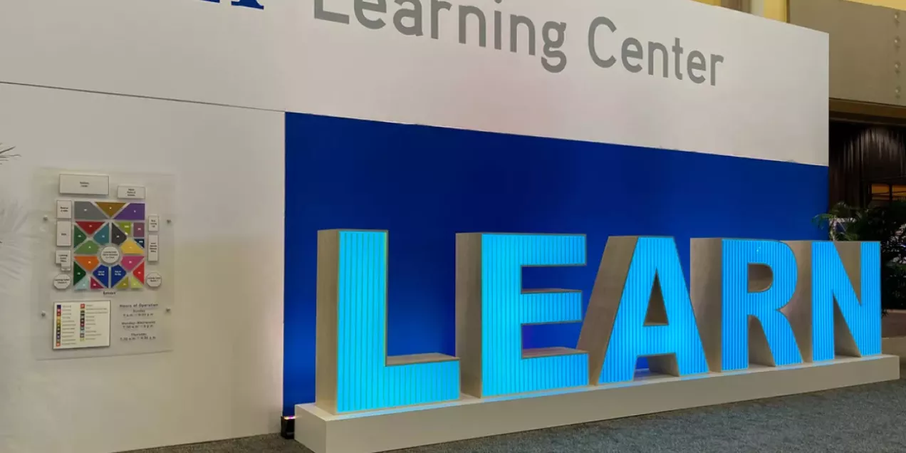 Alt text:  Large freestanding letters spelling “LEARN” displayed as an illuminated exhibit installation. The letters are bright blue with textured vertical detailing and are mounted on a white platform. The display is positioned indoors against a blue-and-white wall, with part of the phrase “Learning Center” visible above. The exhibit appears to be part of a conference, educational, or professional event space designed to promote learning and knowledge sharing.