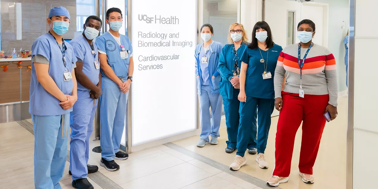 Group of seven masked healthcare professionals standing in a hospital corridor outside UCSF Health Radiology and Biomedical Imaging Cardiovascular Services. Team members wear scrubs, identification badges, and clinical attire while posing together in a bright healthcare setting. A wall sign identifying the cardiovascular imaging department is visible in the background.