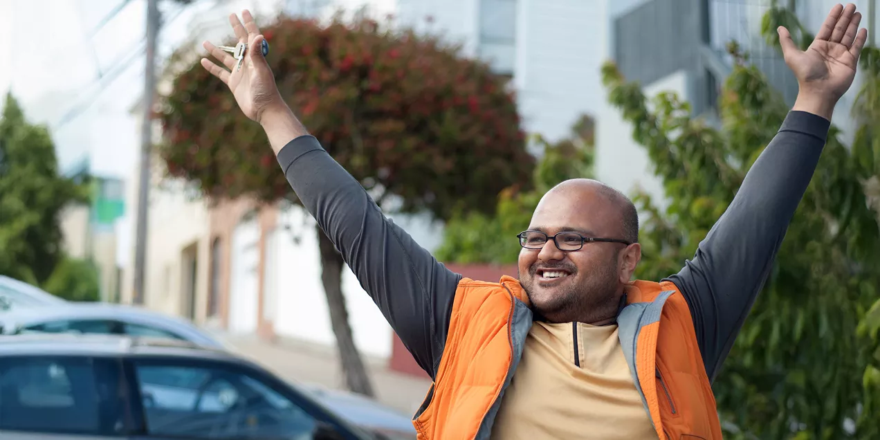 Rahul Desikan smiles and raises his arms while holding keys in one hand, standing on a city street in front of trees and residential buildings.
