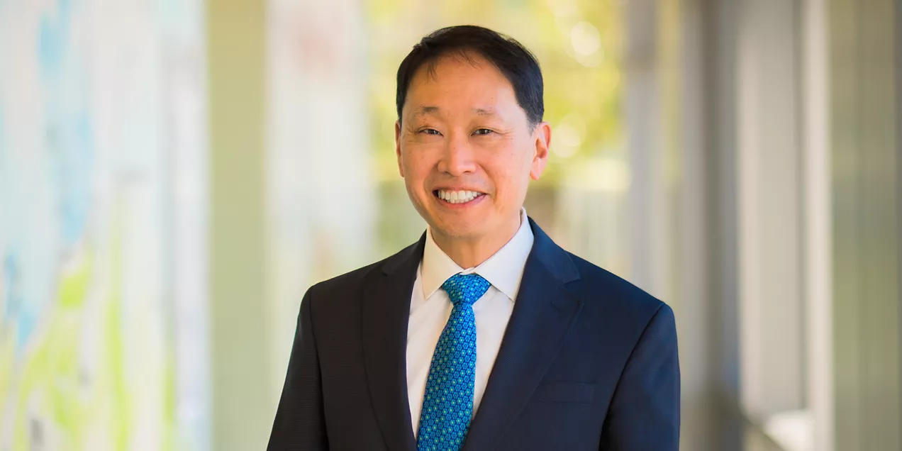 Portrait of a smiling person wearing a dark suit, white shirt, and blue patterned tie. They are standing indoors in a hallway with a softly blurred background, facing the camera.