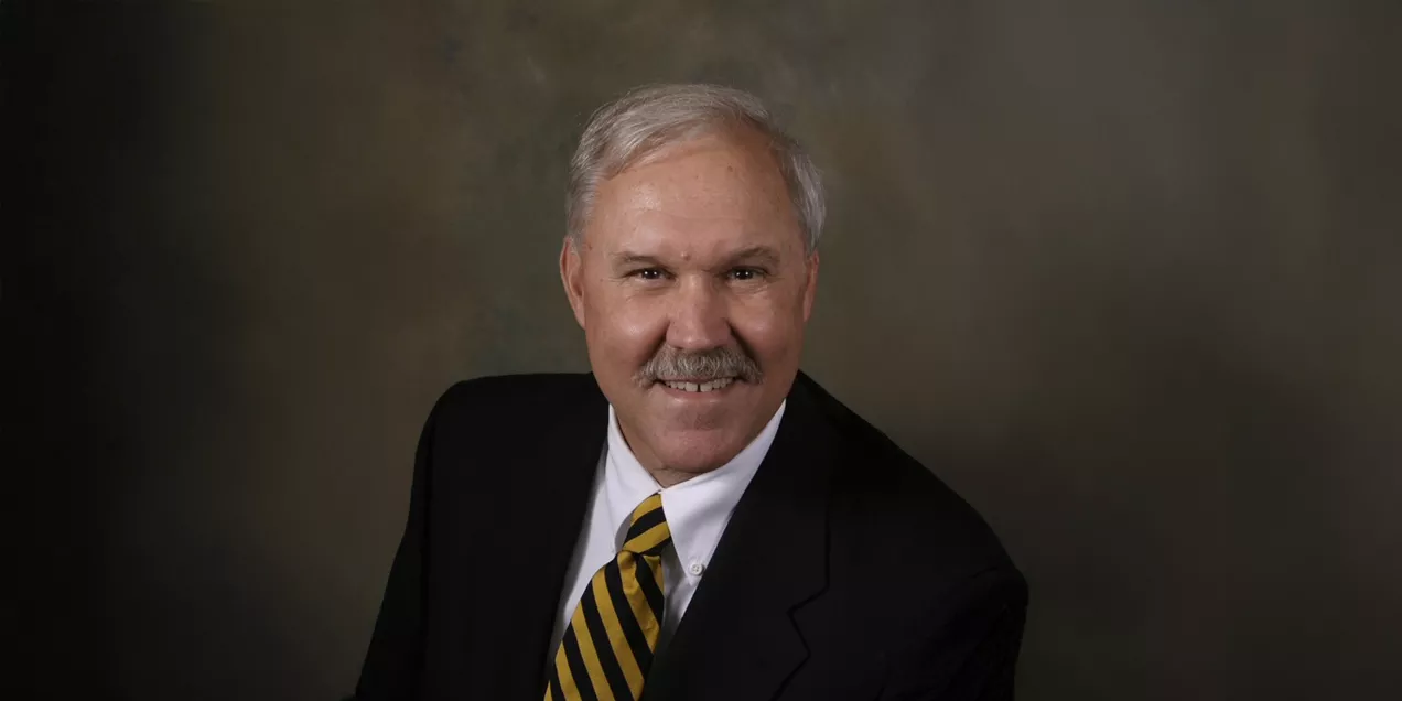 Professional headshot of a man with gray hair and a mustache, wearing a dark suit, white dress shirt, and black-and-gold striped tie. He is smiling at the camera against a softly blended studio background, creating a formal and distinguished portrait.