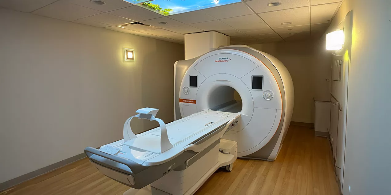 A Siemens Healthineers MRI scanner in a clinical imaging suite with the patient table extended, wood flooring, and soft ambient lighting.