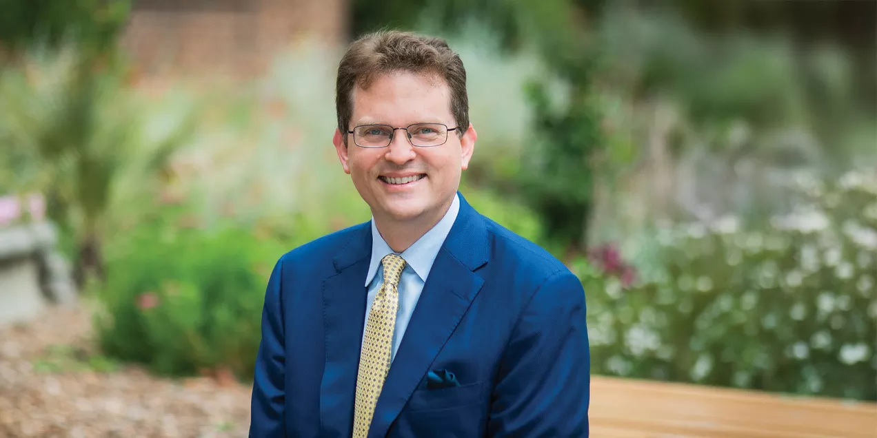 Portrait of a smiling man wearing glasses, a blue suit, and a yellow patterned tie, seated outdoors with a softly blurred garden background.