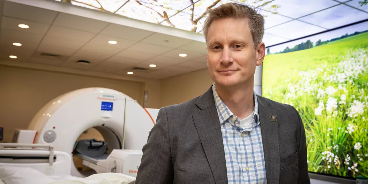 Portrait of a man standing in a modern medical imaging suite beside an MRI scanner. He is wearing a gray blazer over a blue-and-white checkered shirt and is looking directly at the camera with a slight smile. Behind him, the MRI machine and patient table are visible, while large wall and ceiling displays show bright nature scenes with green fields and tree branches. The clinical environment is designed to create a calming atmosphere for patients undergoing imaging procedures.