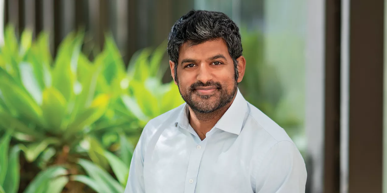 Professional outdoor portrait of a man with short dark hair and a trimmed beard, wearing a light blue button-down shirt. He is seated and smiling at the camera, with lush green plants and a modern building softly blurred in the background. Natural lighting creates a warm and approachable professional headshot.