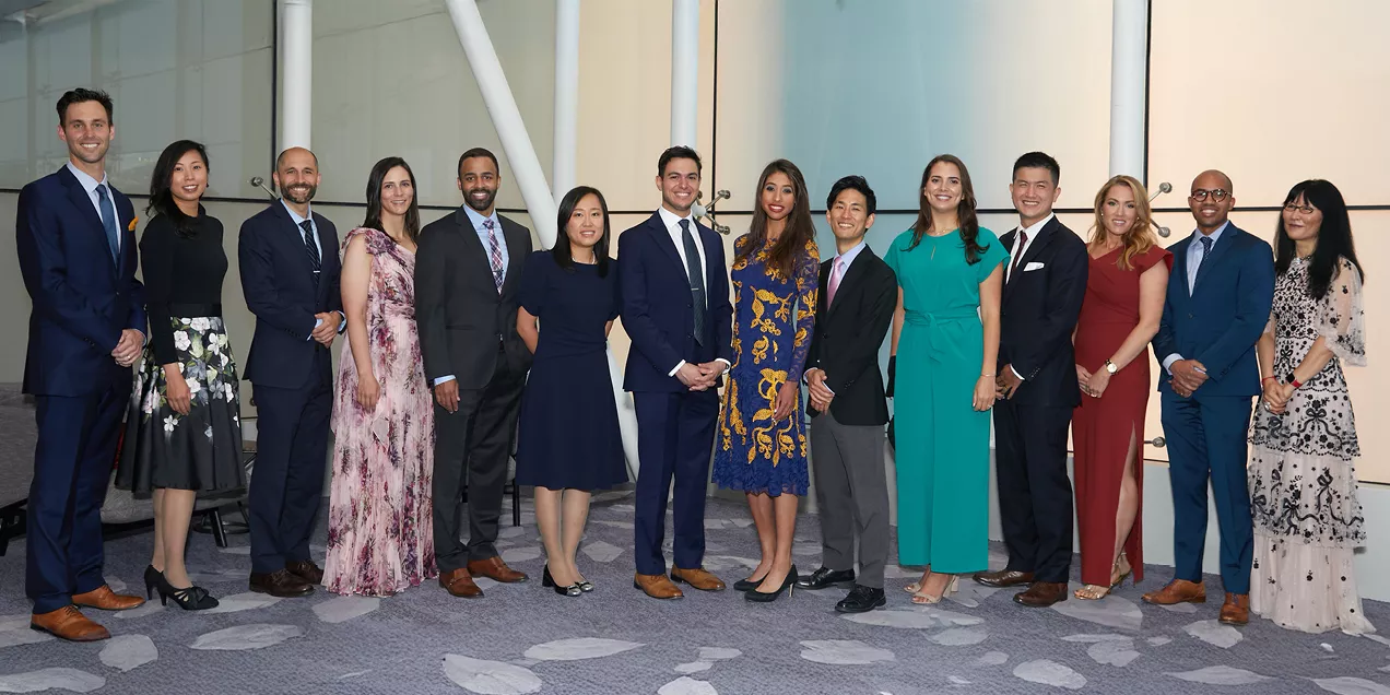 Alt text:  Formal group portrait of fourteen radiology trainees and faculty members standing together at an indoor graduation or awards ceremony. The group is dressed in professional and formal attire, including suits, ties, and dresses in a variety of colors and styles. They are arranged in a single row, smiling toward the camera in a modern event space with glass walls and contemporary architectural features. The image highlights a celebration of academic achievement, professional milestones, and the succ