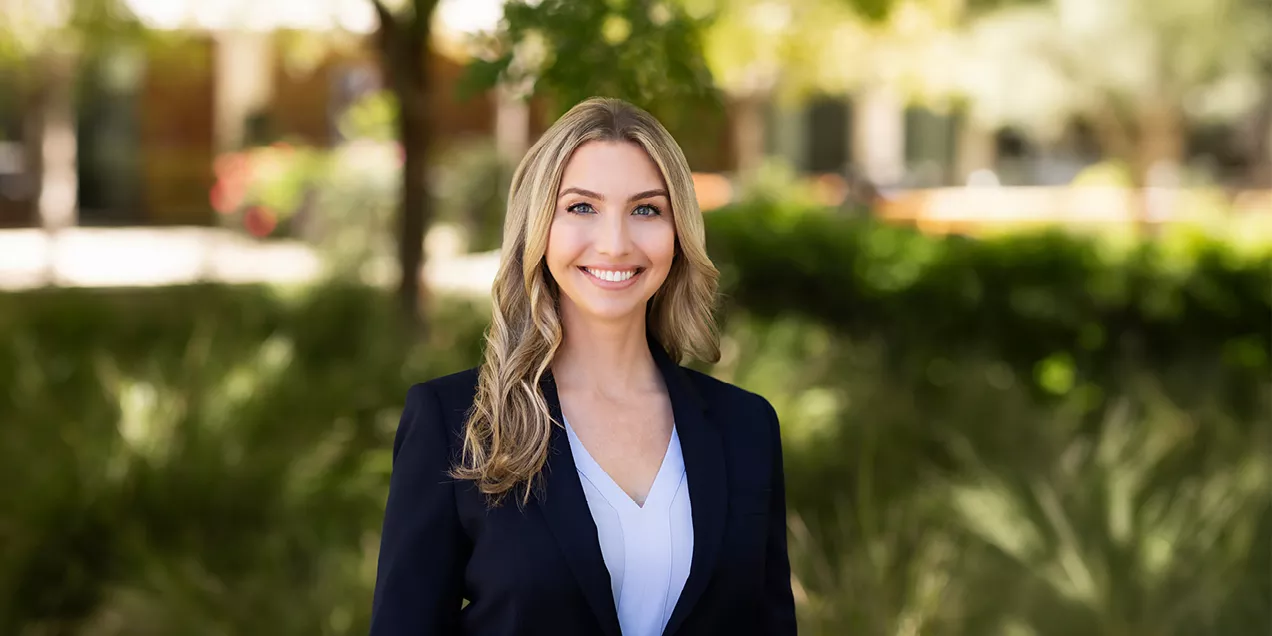 Portrait of a smiling person with long wavy blonde hair wearing a navy blazer over a light blue blouse. They are facing the camera outdoors with a softly blurred background of trees and greenery.
