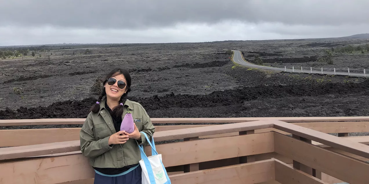 Xiaoxi Liu smiles while standing on a viewing platform in front of a volcanic lava field. Wearing sunglasses and a green jacket, she holds a small purple plush toy and carries a tote bag, with the rugged landscape visible behind her.