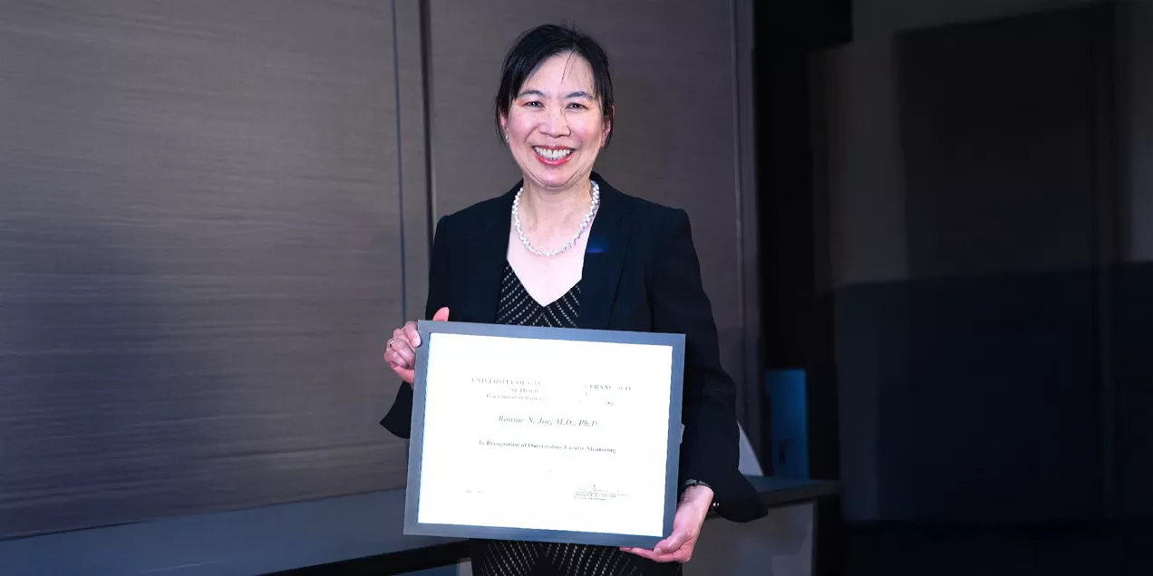 A woman wearing a dark blazer and patterned dress smiles while holding a framed certificate recognizing her outstanding faculty mentoring contributions. She stands indoors at an awards ceremony, wearing a pearl necklace, with the certificate prominently displayed.