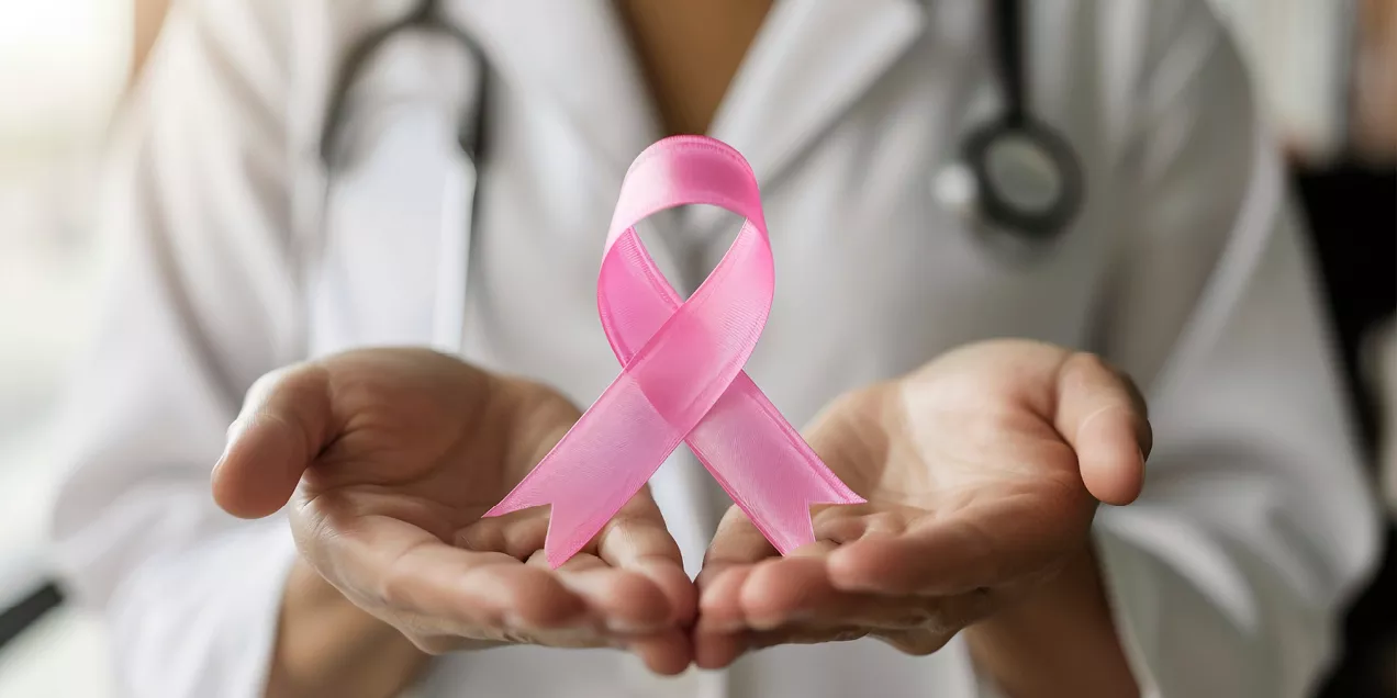 A healthcare professional in a white coat holds a pink breast cancer awareness ribbon in outstretched hands. A stethoscope hangs around their neck, and the softly blurred background keeps the focus on the ribbon, symbolizing support, awareness, and advocacy for breast cancer patients and survivors.