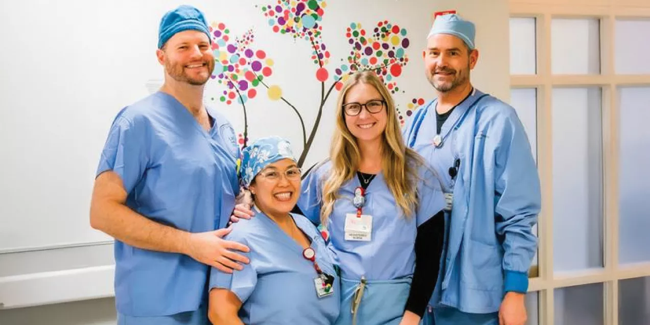 Group photo of four healthcare professionals standing together in a hospital or clinical setting. The team is dressed in blue medical scrubs, with two members also wearing surgical caps. They are smiling toward the camera and standing in front of a colorful wall mural depicting a tree made of multicolored circular leaves. Identification badges are visible on several team members, and one person has a hand resting on a colleague’s shoulder. The image conveys teamwork, compassion, and a supportive healthcare 