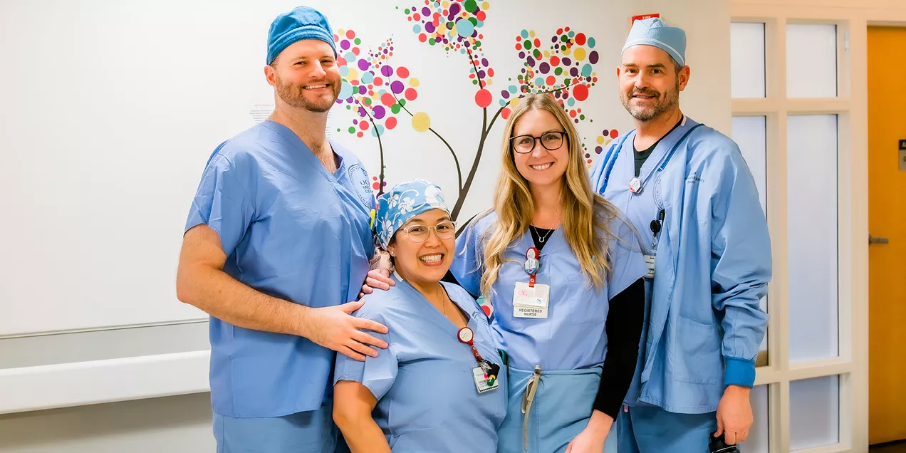 Four healthcare professionals in surgical scrubs stand together smiling in front of a colorful "Gratitude Tree" wall display. The group poses in a hospital hallway, highlighting teamwork, appreciation, and a positive workplace culture.
