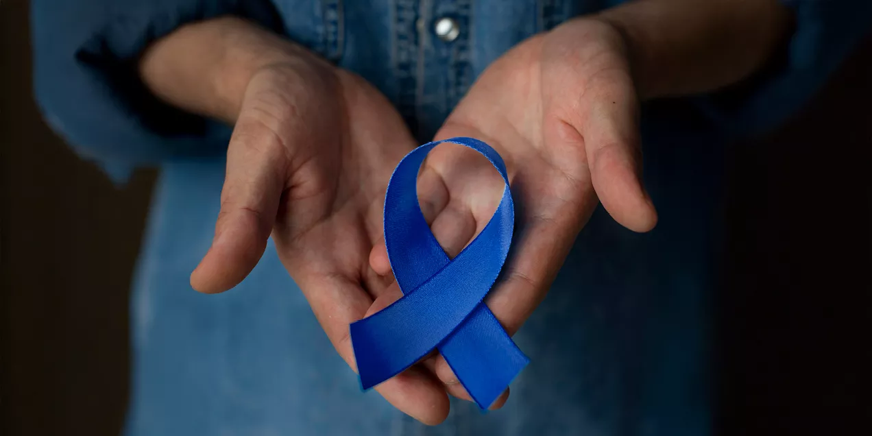 Two open hands gently holding a blue awareness ribbon. The ribbon rests in the center of the palms, symbolizing support and awareness. The background is softly blurred, drawing attention to the ribbon and hands. The blue ribbon is commonly associated with prostate cancer awareness.