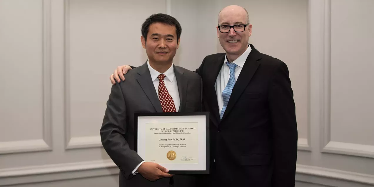 Two people in business attire stand side by side, smiling at the camera. The person on the left holds a framed award certificate while the person on the right has an arm around their shoulder in congratulations. Both are wearing suits and ties, and the photo is taken indoors against a neutral background, marking an award or recognition ceremony.