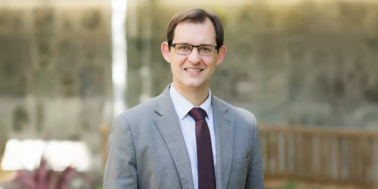 A professionally dressed person wearing a light gray suit, white shirt, and dark burgundy tie smiles at the camera. They have short dark hair and rectangular glasses. The portrait is taken outdoors or in a bright atrium, with a softly blurred background featuring glass panels and a wooden bench.