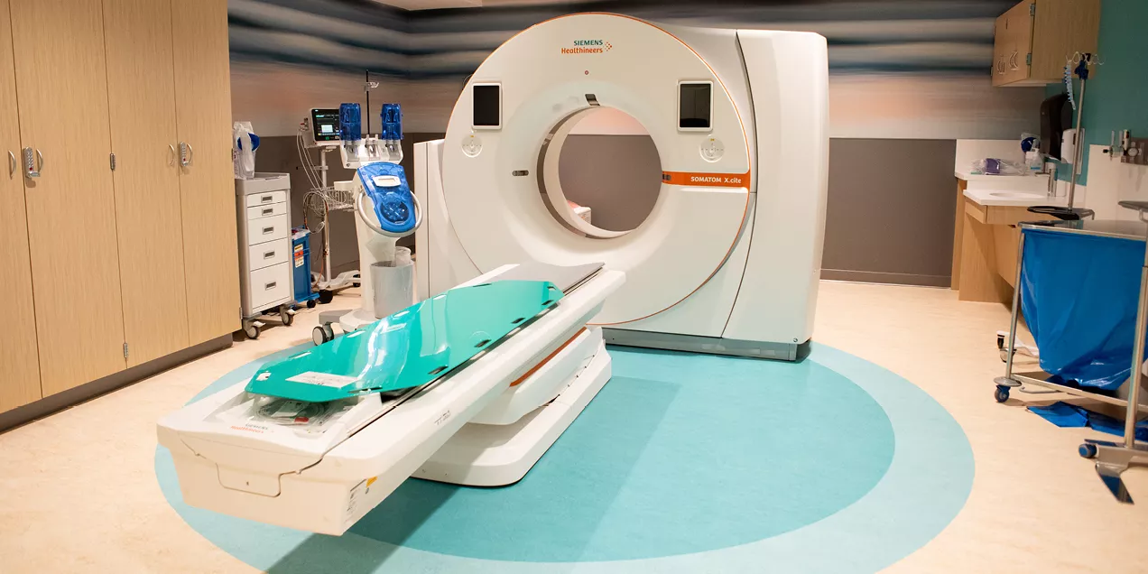 A modern CT imaging suite featuring a large white CT scanner with an attached patient table positioned in the center of the room. Medical equipment, cabinets, and a patient monitoring station surround the scanner, while the clean, brightly lit space has wood cabinetry, teal flooring accents, and a calming striped wall design.