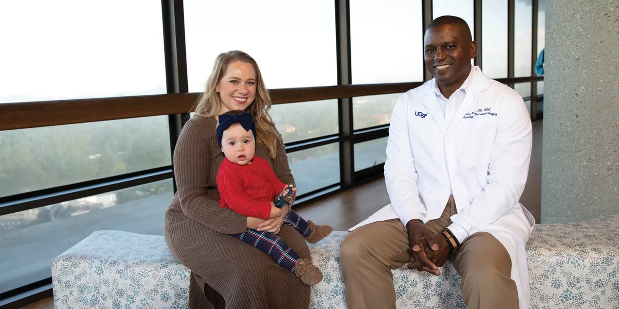 UCSF radiologist Kevin McGill sits with patient Maryana Kessel and her young child in a hospital corridor overlooking large windows. Kessel holds her child on her lap while McGill, wearing a white medical coat, sits beside them. The image highlights compassionate, family-centered care in a clinical setting.