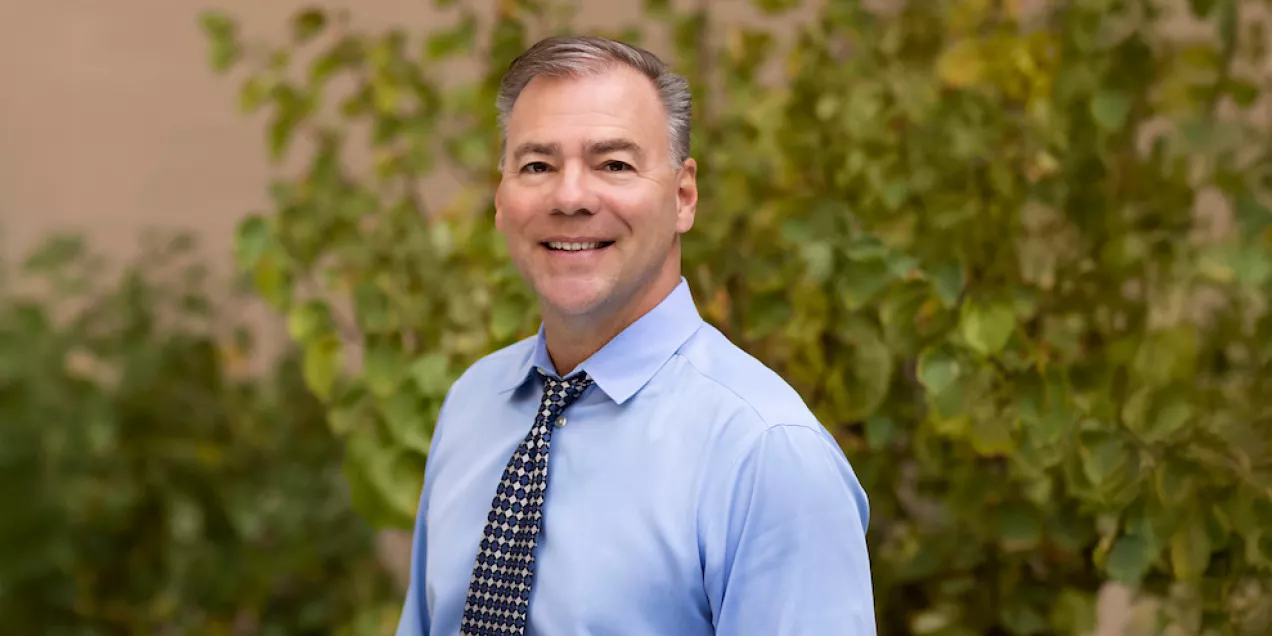 Professional headshot of a smiling person with short gray hair wearing a light blue dress shirt and a patterned navy tie. The portrait is taken outdoors or in a garden setting, with green foliage softly blurred in the background.