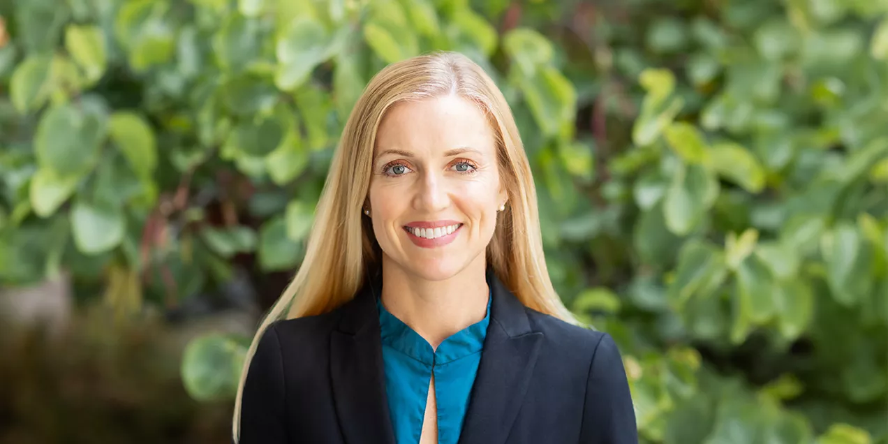 Portrait of Brittany Harrison smiling, wearing a black blazer over a teal blouse, standing outdoors in front of a blurred green foliage background.