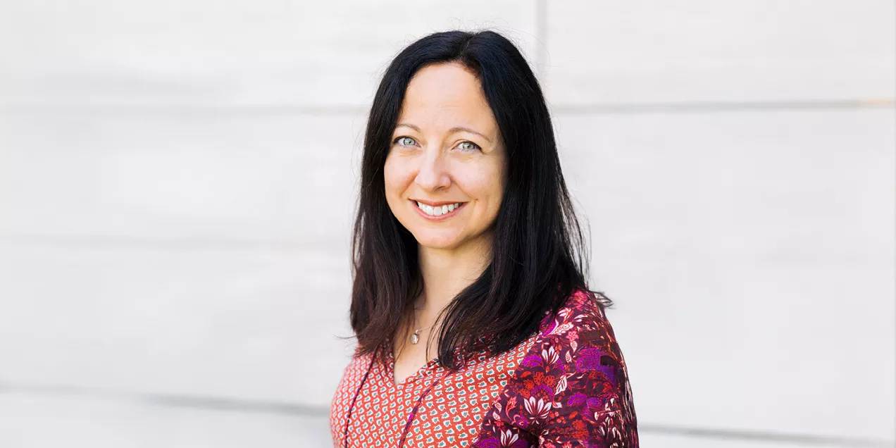 Janine Lupo smiles at the camera in a professional headshot, wearing a patterned red and pink blouse against a light stone background.