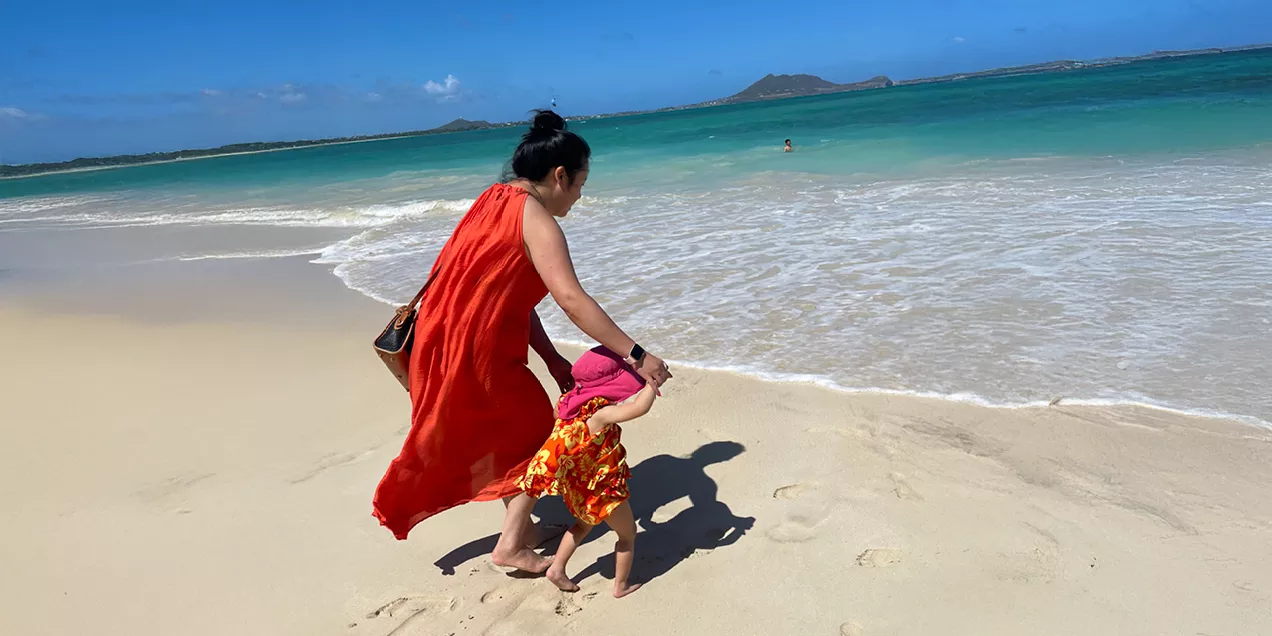 A woman in a flowing red dress holds the hand of a young child wearing a pink sun hat and colorful outfit as they walk barefoot along a sandy beach. Turquoise ocean water, gentle waves, and distant islands stretch beneath a bright blue sky.