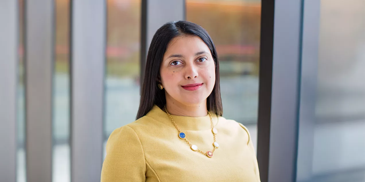 A professional head-and-shoulders portrait of a person standing indoors in front of large floor-to-ceiling windows. She is wearing a light yellow long-sleeve top, small gold earrings, and a gold necklace with blue, white, and red circular accents. She faces the camera with a slight smile. The softly blurred background shows glass panels and an outdoor landscape, creating a bright, modern office or campus setting.