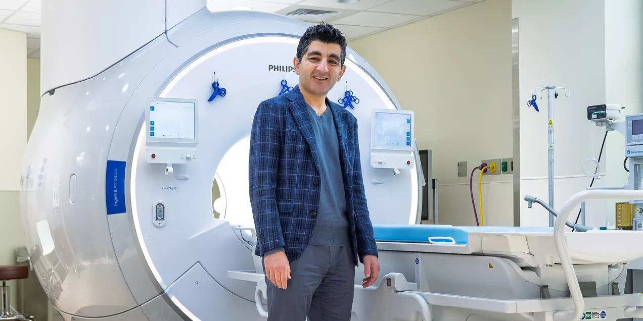Professional portrait of a smiling man wearing a blue plaid blazer over a blue shirt, standing in front of an MRI scanner in a medical imaging suite. The scanner and its control displays are visible in the background.
