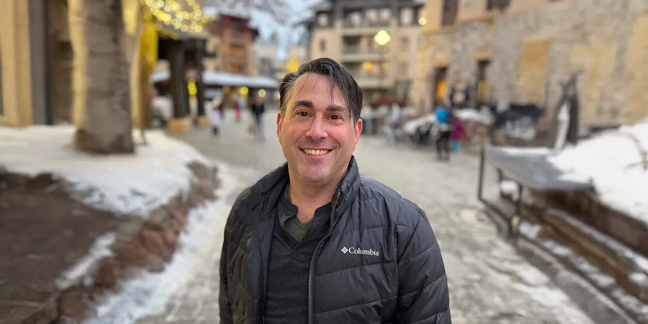 Professional portrait of a smiling man wearing a black jacket over a dark shirt, standing on a snowy pedestrian street with shops and buildings softly blurred in the background.