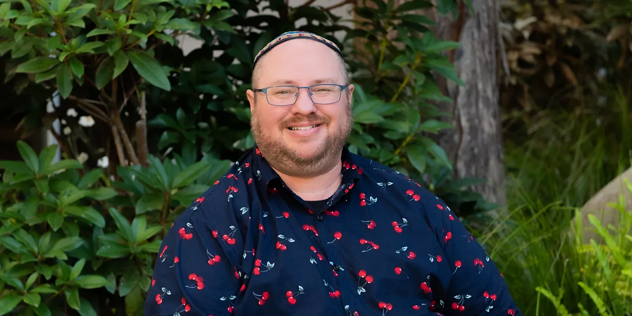 A professional head-and-shoulders portrait of a smiling person seated outdoors in front of lush green foliage. He is wearing glasses, a dark button-up shirt with a red cherry pattern, and a colorful skullcap. He is facing the camera with a friendly expression in a natural, softly blurred setting.
