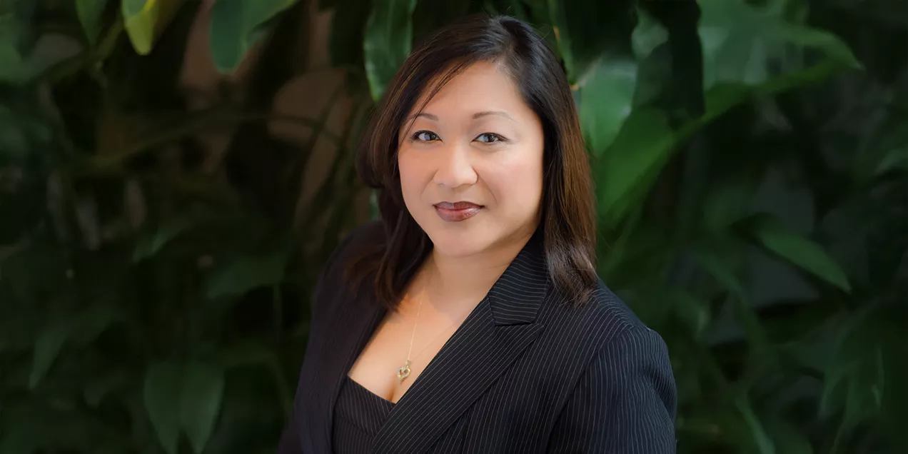A professional head-and-shoulders portrait of a person wearing a dark pinstripe blazer and matching top. She is facing the camera with a slight smile, wearing a delicate necklace, and standing in front of softly blurred green foliage in an indoor setting.
