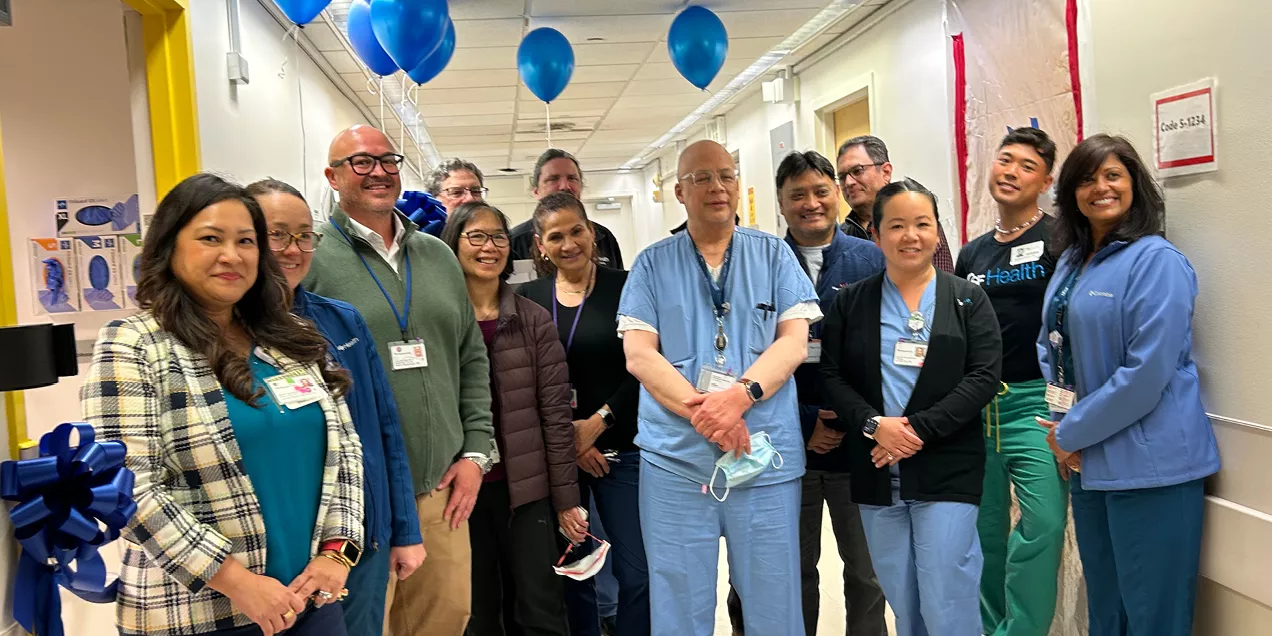 A group of healthcare staff pose together in a hospital hallway during a ribbon-cutting celebration. Blue balloons and smiles mark the occasion, with clinicians and staff wearing a mix of scrubs, lab coats, and business attire.