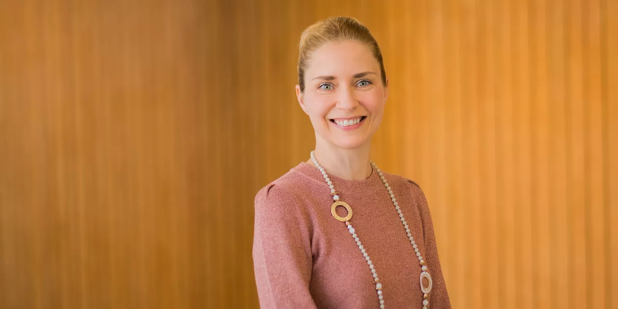 A professional head-and-shoulders portrait of a smiling person standing in front of a warm-toned wood-paneled background. She is wearing a rose-colored long-sleeve top and a long beaded necklace with circular accents. Her hair is pulled back, and she is facing the camera with a friendly expression.