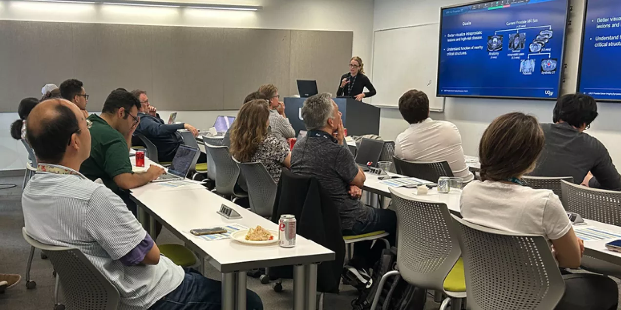 A presenter stands at the front of a conference room delivering a presentation to an audience seated at tables. Attendees listen while viewing a large screen displaying slides about prostate cancer imaging and artificial intelligence. Laptops, notebooks, and refreshments are visible, highlighting an educational symposium or professional training session.