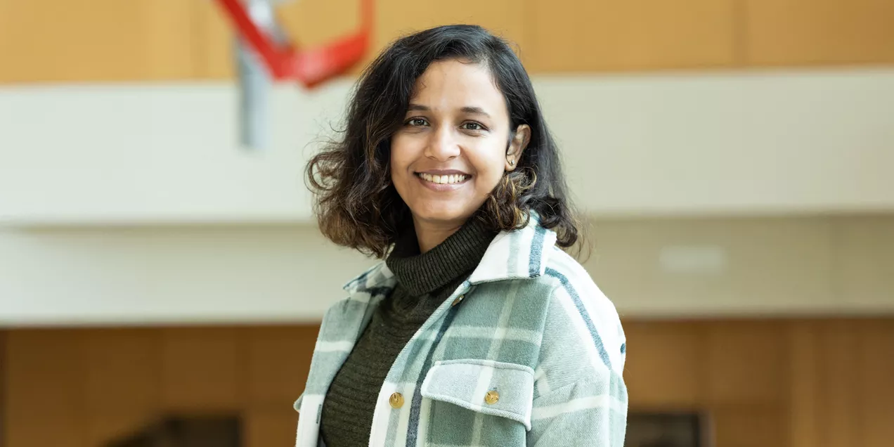 Rupsa Bhattacharjee smiling in an indoor portrait, wearing a green turtleneck and a light plaid overshirt.