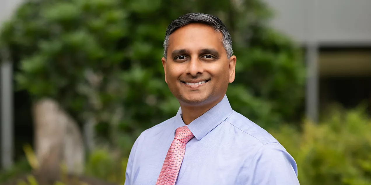 Professional headshot of a smiling man wearing a light blue dress shirt and a pink patterned tie, standing outdoors with blurred green foliage in the background.