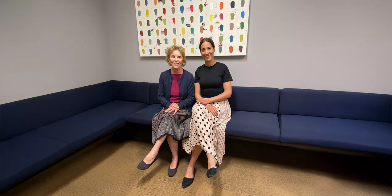 Two women sit together on a blue bench beneath a colorful abstract painting, smiling at the camera.